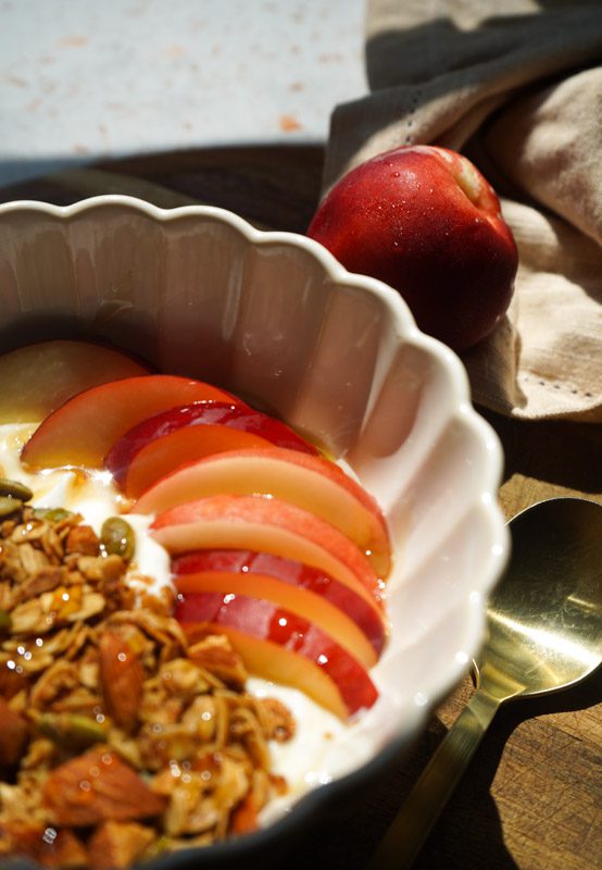 Angled view of honey nectarine yogurt bowl in white scalloped bowl with gold spoon fresh nectarine slices honey roasted granola honey drizzle and whole nectarine in linen background