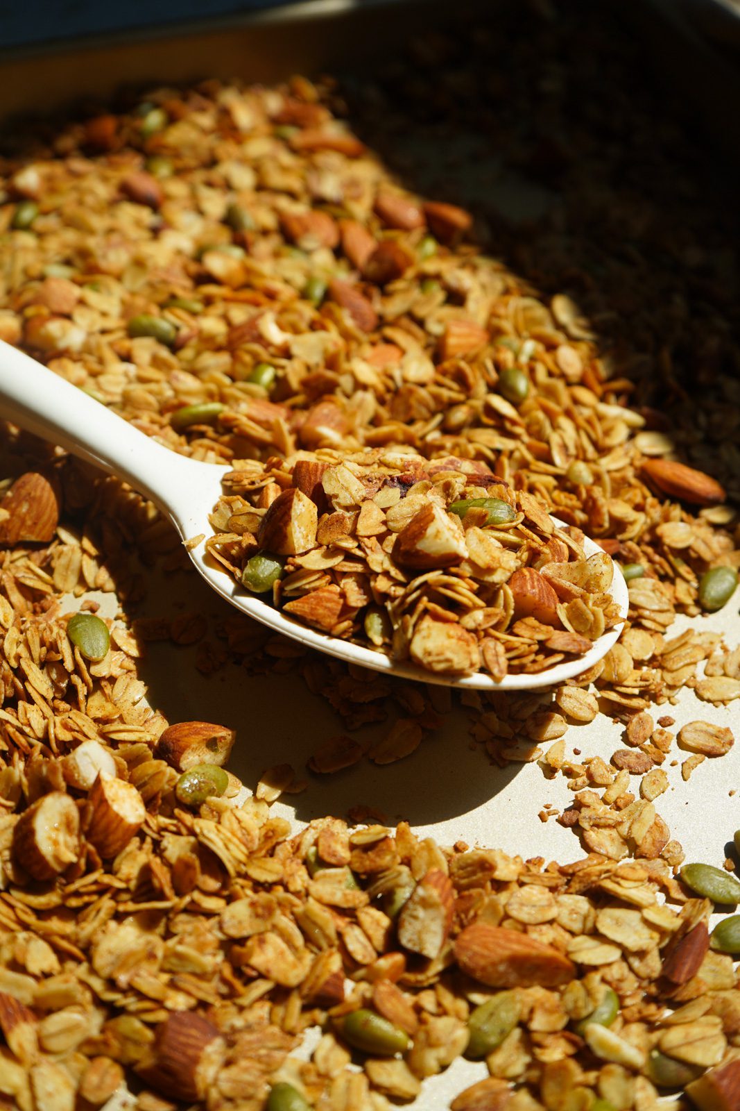 Spoon scooping homemade honey roasted almond granola from a baking sheet with oats, almonds, and pumpkin seeds.