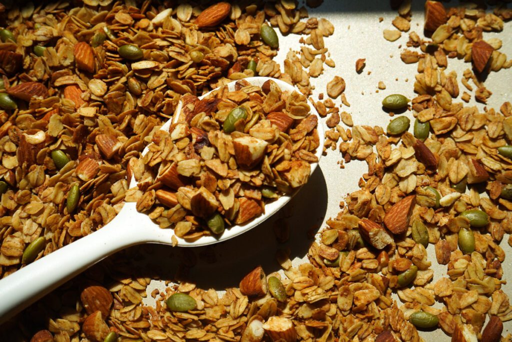 Spoon scooping homemade honey roasted almond granola from a baking sheet with oats, almonds, and pumpkin seeds.