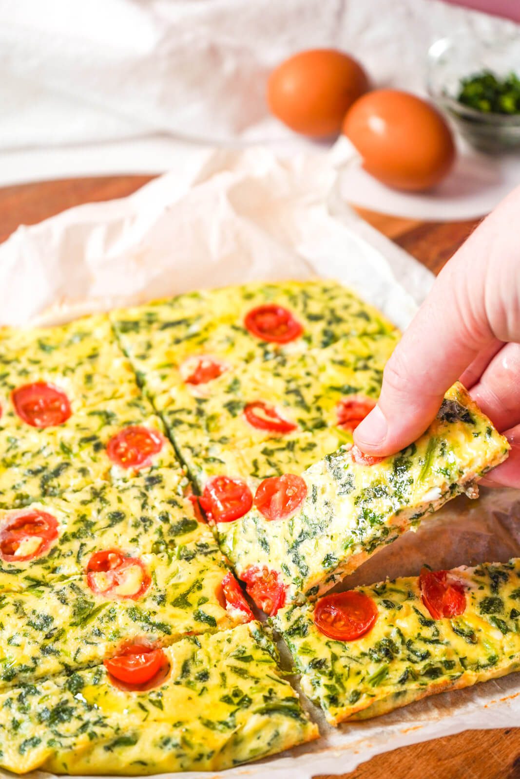 Savory egg and spinach fingers with tomato slices held up to show detail over a wooden board with eggs in the background.