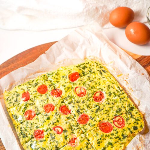 Baked protein egg and spinach fingers with cherry tomato arranged on parchment paper over a wooden board with fresh eggs nearby.