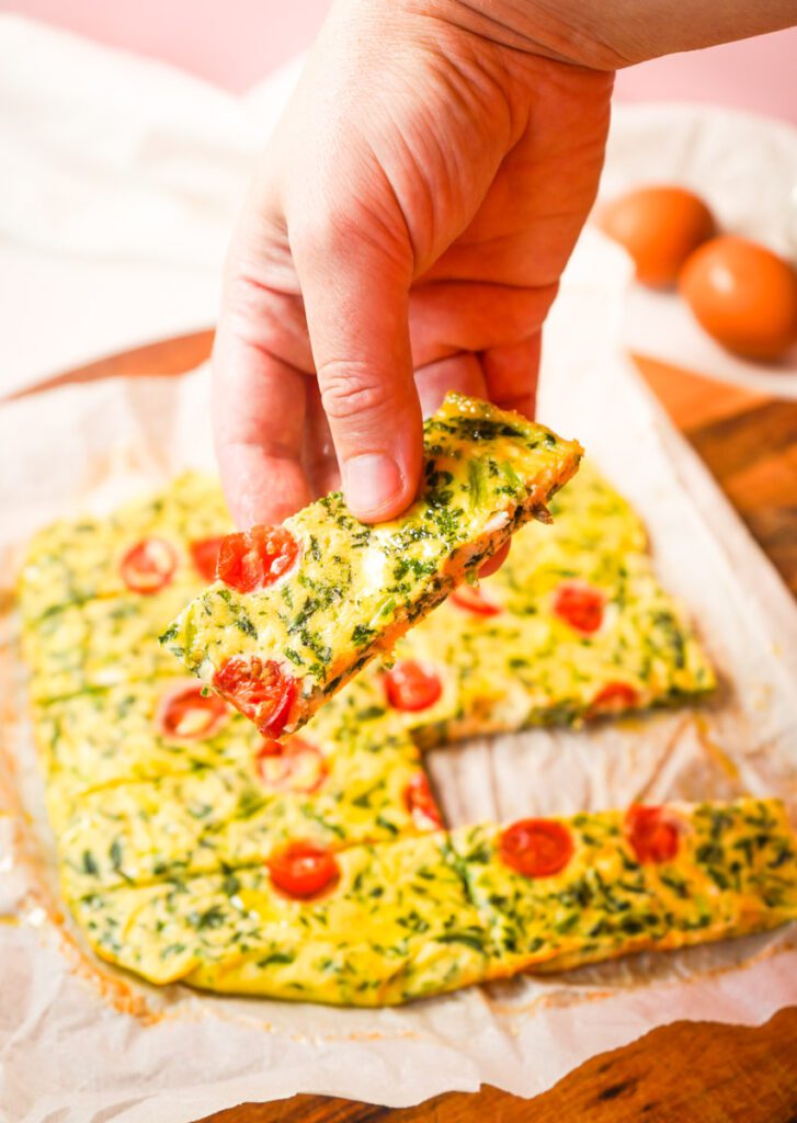 Mom's hand holding a baked egg and spinach finger with cherry tomato, showing the firm, custard-like texture perfect for little hands.