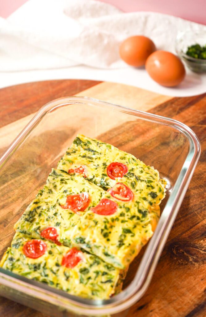 Baked egg and spinach fingers stored in a glass container on a wooden board, ready for meal prep and toddler snacking.