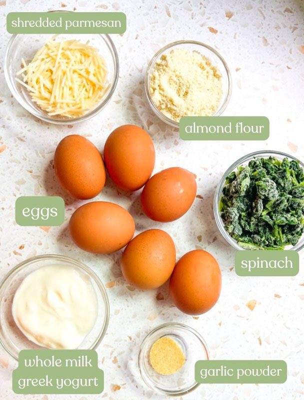 Ingredients for protein egg and spinach fingers: eggs, shredded Parmesan, almond flour, Greek yogurt, fresh spinach, and salt arranged in glass bowls on a marble counter.