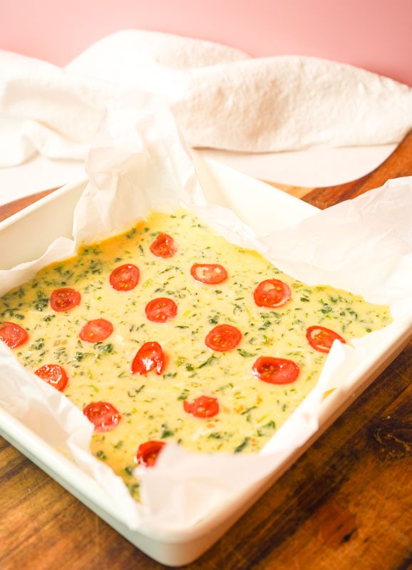Protein egg and spinach fingers with tomato on parchment paper, displayed on a wooden board against a soft pink background.