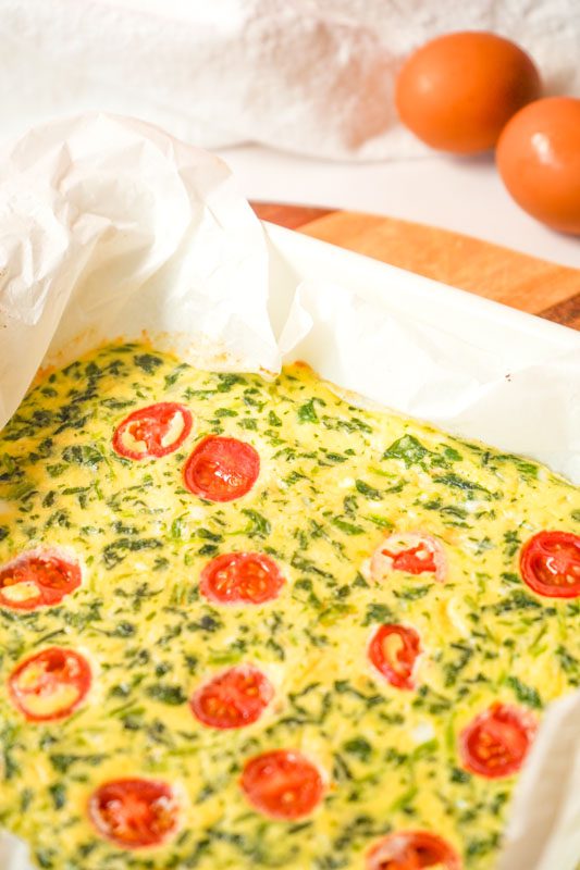 Baked protein egg and spinach fingers with cherry tomato in a white ceramic baking dish lined with parchment, ready to cool.