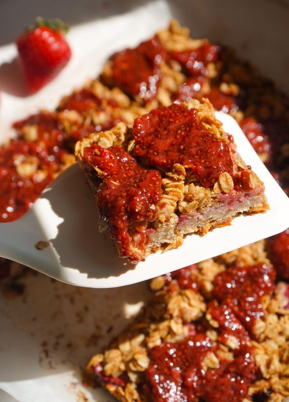 Strawberry chia jam oat squares lifted on a white spatula showing golden oat crumble and deep ruby strawberry chia jam on top