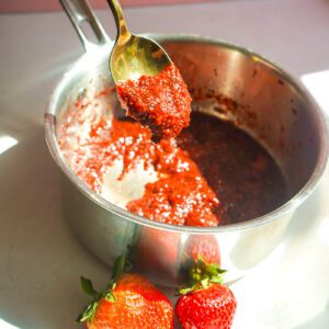A gold spoon scooping thick homemade strawberry chia seed jam out of a stainless steel saucepan with fresh strawberries nearby.