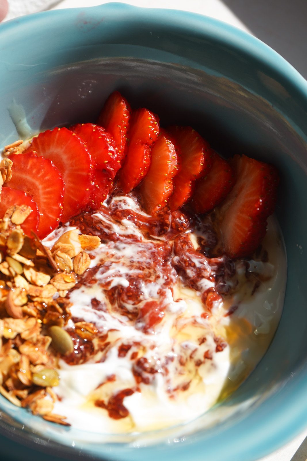 Close up overhead view of strawberry chia jam swirled through white Greek yogurt in teal bowl with fresh sliced strawberries and homemade honey almond granola in warm natural light