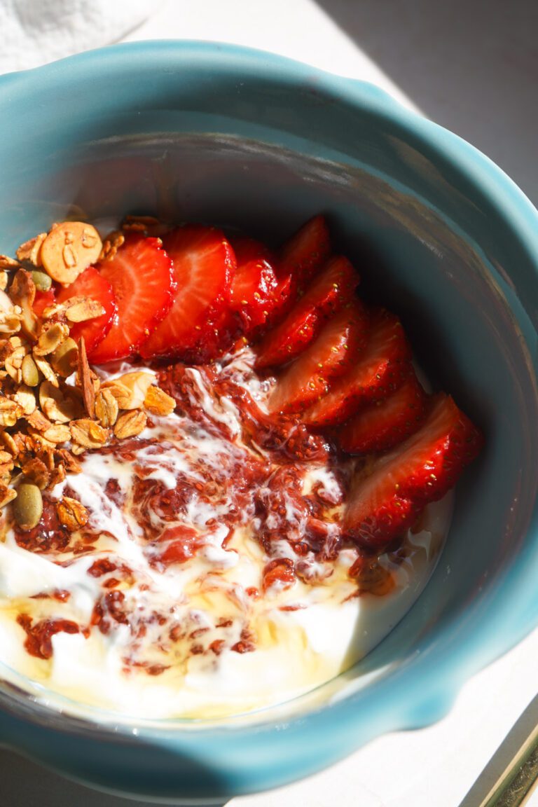 Close up overhead view of strawberry chia jam swirled through white Greek yogurt in teal bowl with fresh sliced strawberries and homemade honey almond granola in warm natural light
