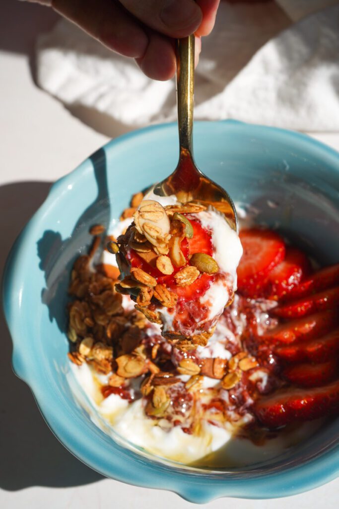 Hand lifting a gold spoon loaded with yogurt granola and strawberry chia jam from a teal bowl showing the perfect bite with fresh strawberries in background