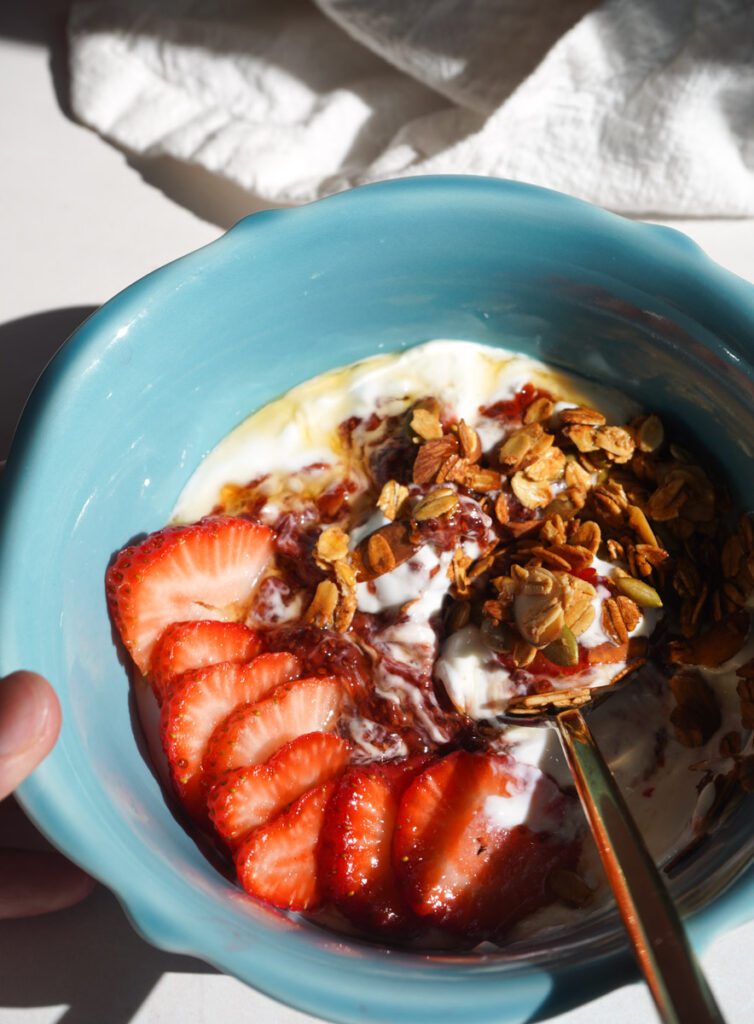 Close up overhead view of strawberry chia jam swirled through white Greek yogurt in teal bowl with fresh sliced strawberries and homemade honey almond granola in warm natural light