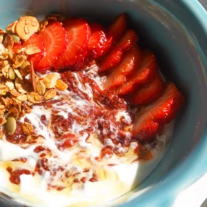 Close-up overhead shot of a blue ceramic bowl with swirled yogurt, strawberry chia jam, fresh sliced strawberries, crunchy granola, and pumpkin seeds.