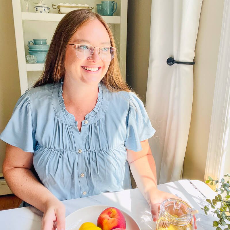 Amy smiling in her kitchen wearing a light blue top, holding a jar in front of a sunlit window.