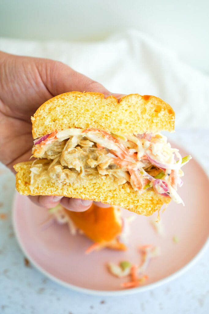 Close-up of a hand holding a cut apple cider pulled chicken sandwich showing juicy shredded chicken and creamy apple slaw.