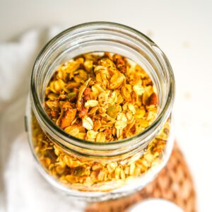 Overhead close-up of freshly baked apple cinnamon pecan granola with oats, pecans, pepitas, and baked apples in a glass jar.