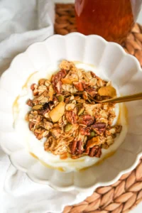A white ceramic bowl filled with thick Greek yogurt topped with crunchy apple cinnamon pecan granola and a honey drizzle.