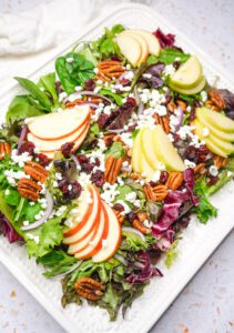 A gorgeous seasonal apple pecan salad served on a white platter, garnished with goat cheese, pecans, dried cranberries, and red onion.