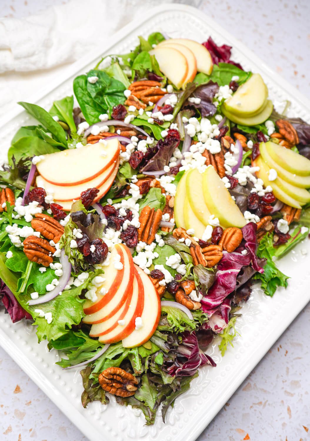 A gorgeous seasonal apple pecan salad served on a white platter, garnished with goat cheese, pecans, dried cranberries, and red onion.