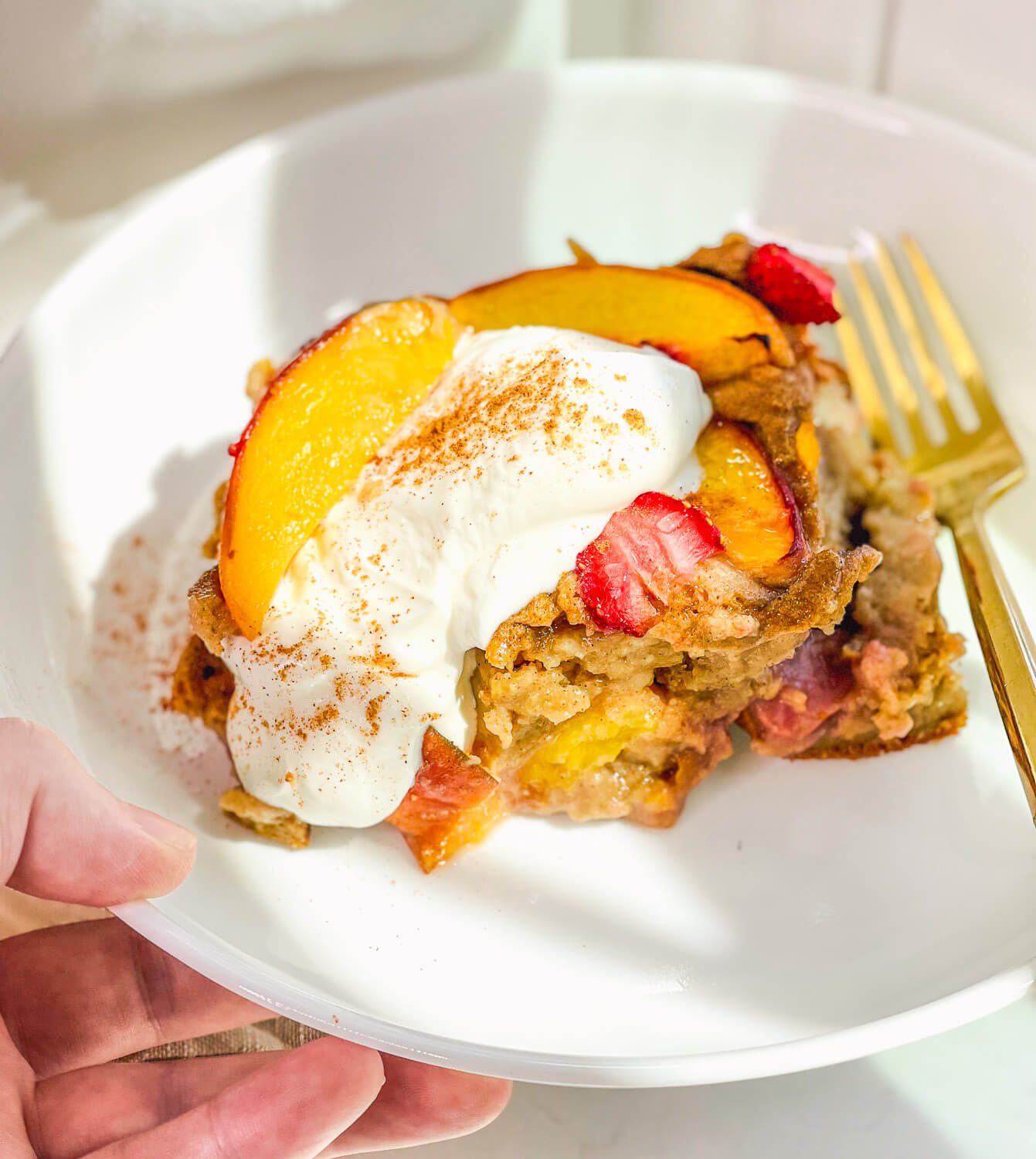 A person holding a white plate with a slice of strawberry peach baked oatmeal under bright morning sunlight.