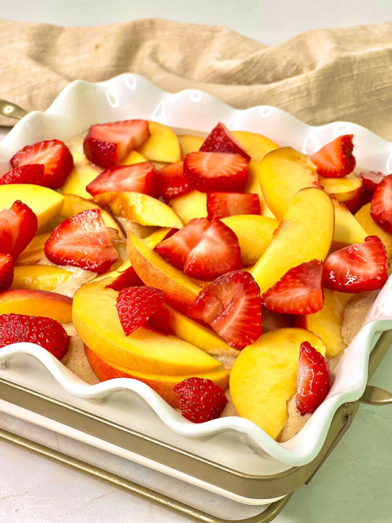 Unbaked high-protein strawberry peach oatmeal batter in a white ceramic dish topped with fresh fruit.