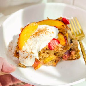 Close-up shot of a warm square of strawberry peach baked oatmeal topped with a dollop of white Greek yogurt and cinnamon on a white plate.