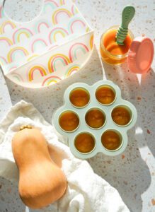 Green silicone freezer tray filled with bright orange butternut squash puree alongside a glass jar and silicone spoon.