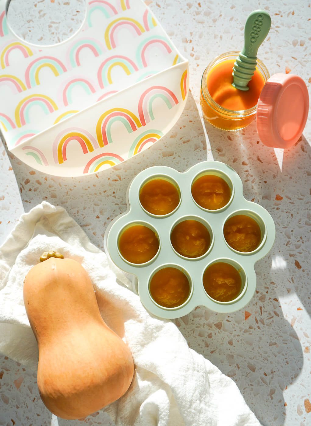 Green silicone freezer tray filled with bright orange butternut squash puree alongside a glass jar and silicone spoon.