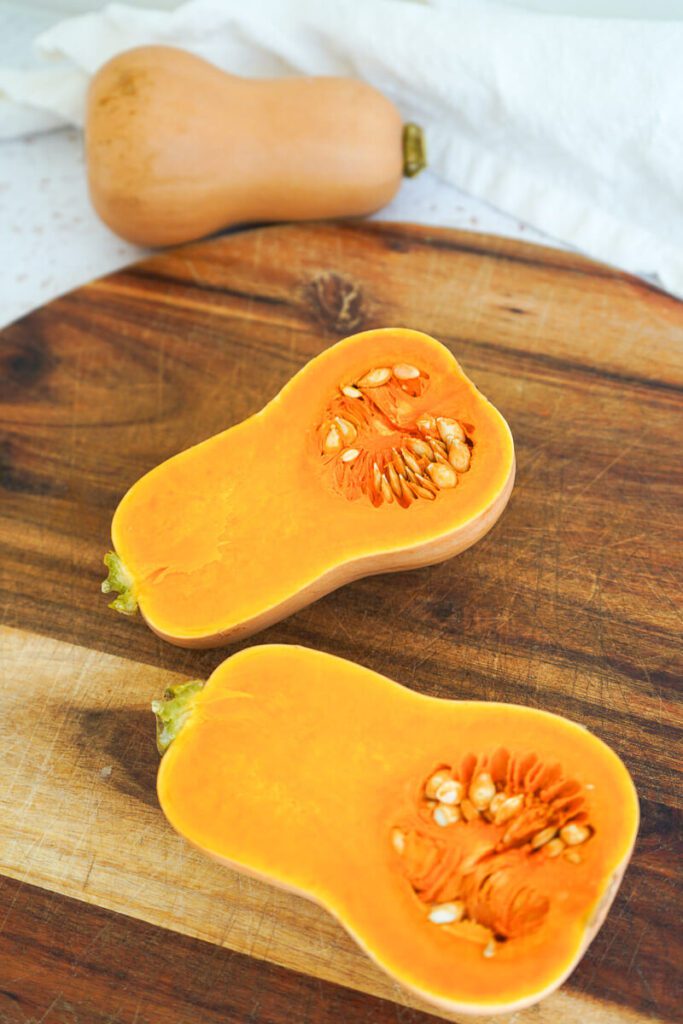 Two butternut squash halves resting on wooden cutting board, whole squash in background with linen napkin.