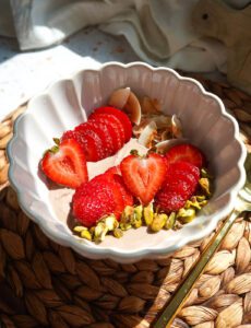 Scalloped ceramic bowl filled with dark chocolate Greek yogurt topped with fresh sliced strawberries, toasted coconut chips, and green pistachios.