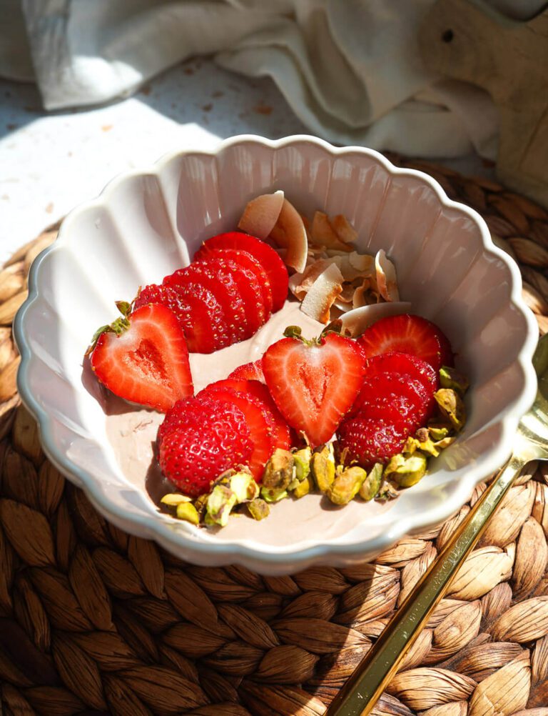 Scalloped ceramic bowl filled with dark chocolate Greek yogurt topped with fresh sliced strawberries, toasted coconut chips, and green pistachios.