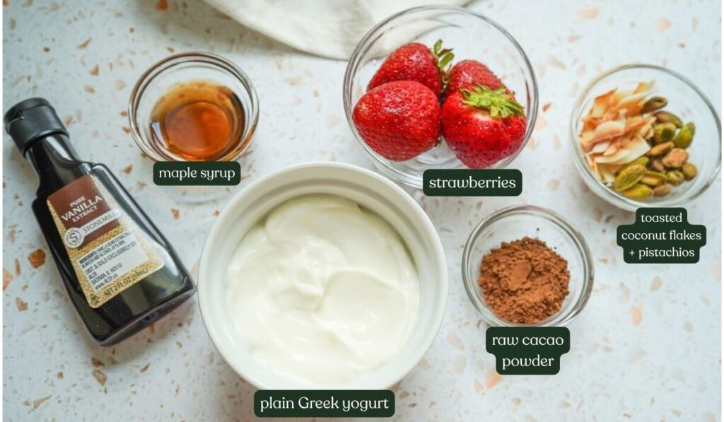 Plain Greek yogurt, raw cacao powder, pure maple syrup, fresh strawberries, coconut, and pistachios on a white terazzo counter.