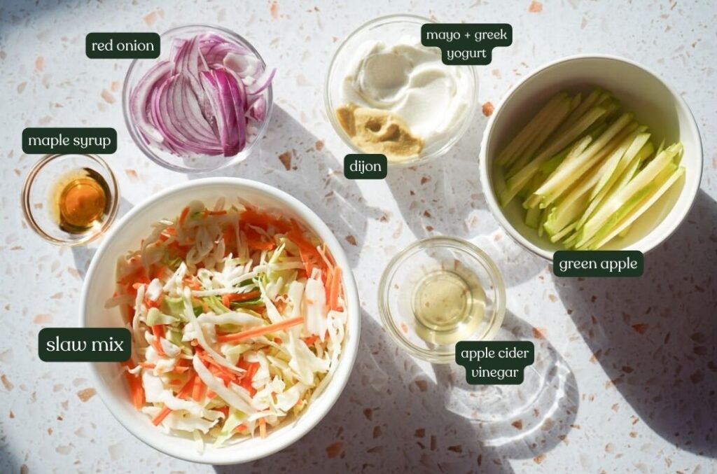 Fresh ingredients for creamy apple slaw including coleslaw mix, Granny Smith apples, red onion, mayo, Greek yogurt, and Dijon mustard.