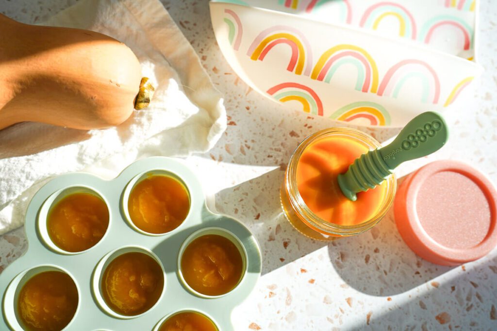 Green silicone freezer tray filled with bright orange butternut squash puree alongside a glass jar and silicone spoon, ready for serving.