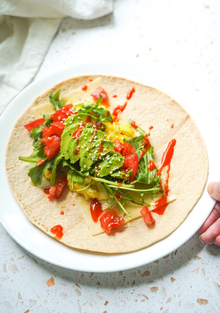 Plate showing assembly of open whole wheat wrap layered with swiss cheese slices, fluffy scrambled egg, arugula, avocado and hot sauce, ready to be rolled up.