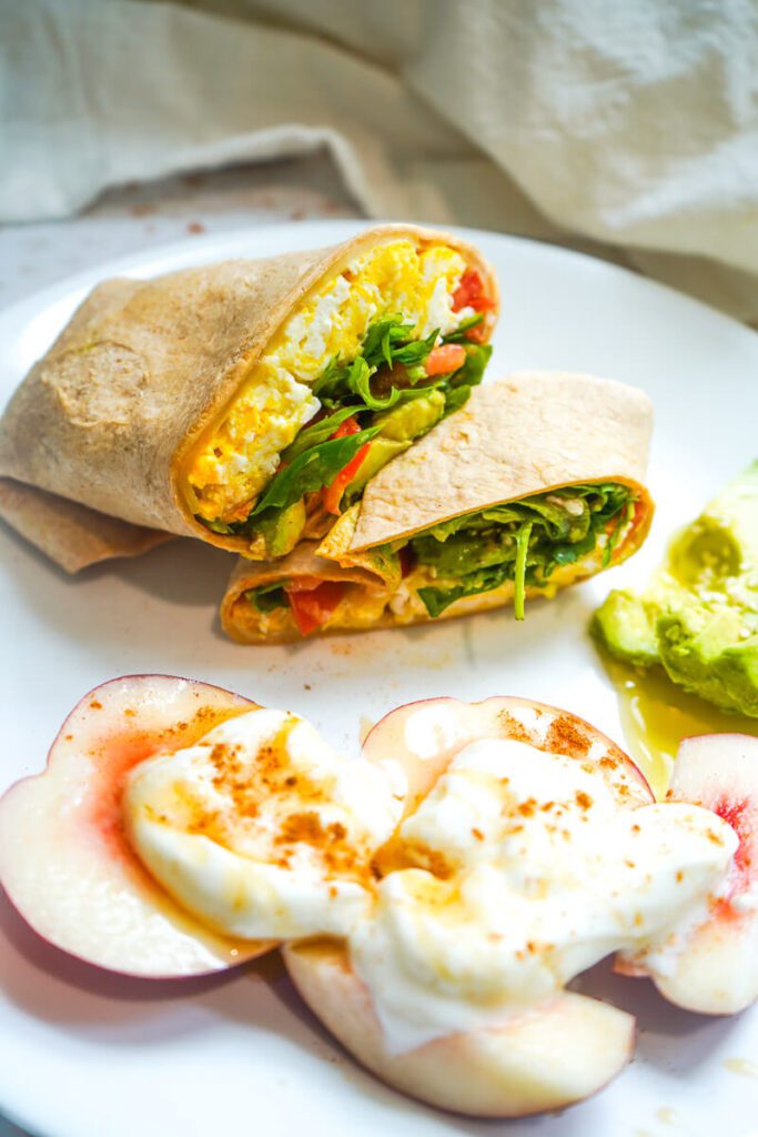 A whole wheat scrambled egg wrap sliced diagonally on a plate next to fresh sliced nectarines topped with cinnamon Greek yogurt and sliced avocado.