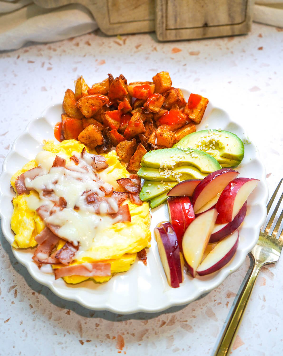 Overhead view of a white ceramic plate filled with cheesy scrambled eggs with ham, crispy air-fryer potatoes, fresh avocado slices with sesame seeds, and juicy nectarine wedges.