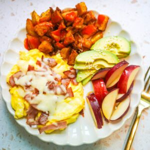 Overhead flat lay of a complete ham and swiss breakfast plate with eggs, potatoes, avocado, and nectarines on a white scalloped plate.