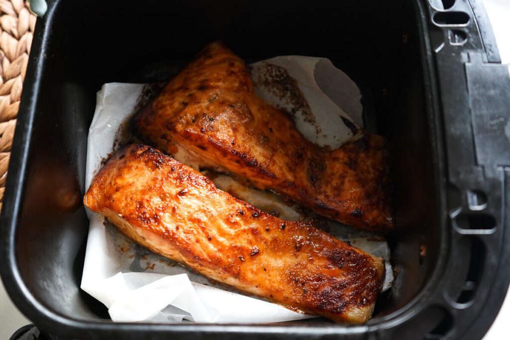 Two golden caramelized salmon fillets cooked inside a black air fryer basket on parchment paper.