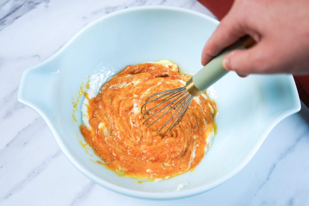 Hand whisking pumpkin puree, Greek yogurt, maple syrup, and eggs in a light blue mixing bowl