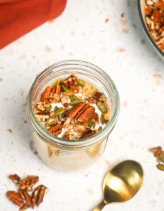 Close-up overhead view of a glass mason jar filled with creamy pumpkin overnight oats, topped with a dollop of Greek yogurt and maple-toasted pecans and pepitas, alongside a gold spoon on a white marble countertop.