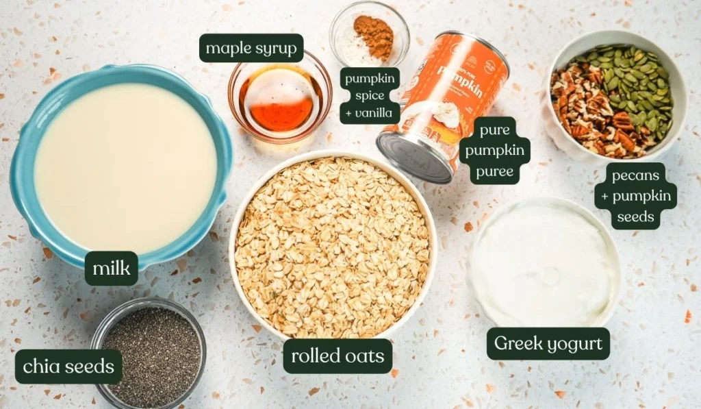 Ingredient flat lay on a white speckled countertop featuring rolled oats in a bowl, a can of pure pumpkin puree, Greek yogurt, milk, maple syrup, chia seeds, and bowls of pecans and pepitas.