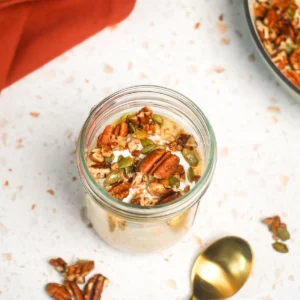 Close-up overhead view of a glass mason jar filled with creamy pumpkin overnight oats, topped with a dollop of Greek yogurt and maple-toasted pecans and pepitas, alongside a gold spoon on a white marble countertop.