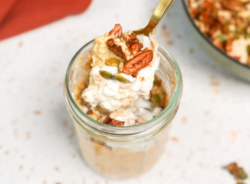 Close-up spoon dipping into an open glass jar of creamy pumpkin overnight oats topped with a yogurt dollop and maple-toasted pecans.