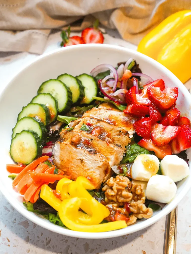 A vibrant bistro-style strawberry balsamic grilled chicken salad in a white bowl with fresh strawberries, cucumbers, and mozzarella pearls.