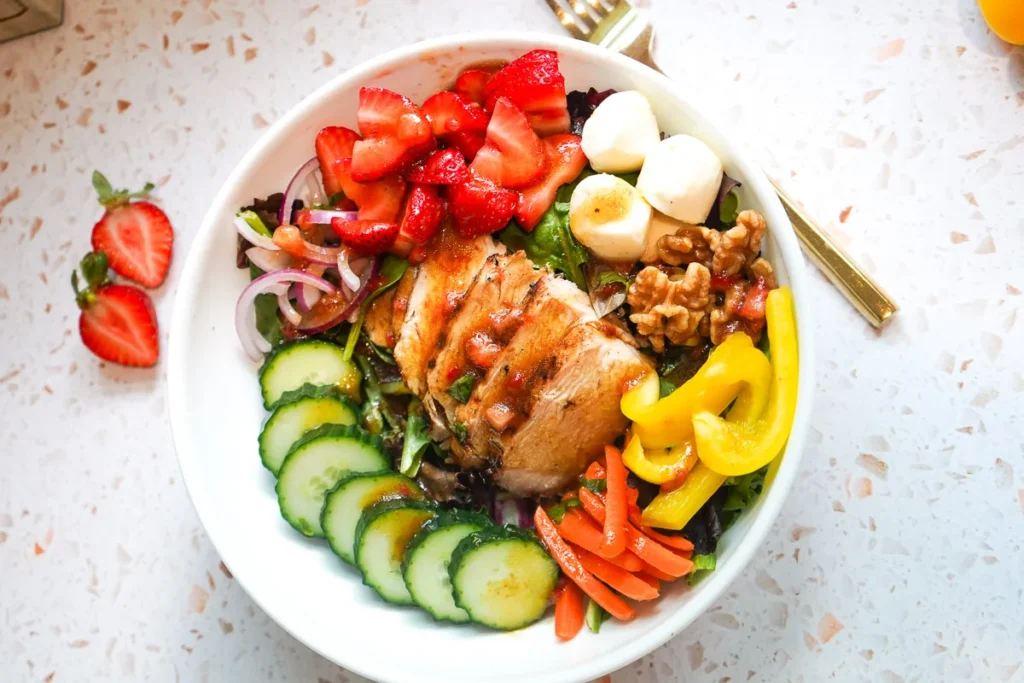 Overhead view of a strawberry balsamic grilled chicken salad bowl loaded with golden sliced chicken, fresh strawberries, cucumbers, yellow bell peppers, red onion, walnuts, and mozzarella pearls.