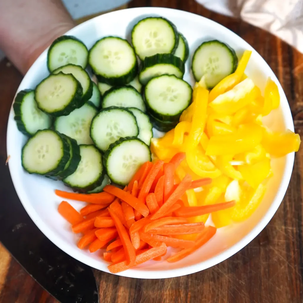 A round white plate filled with sliced English cucumbers, sliced yellow bell peppers, and shredded carrots.