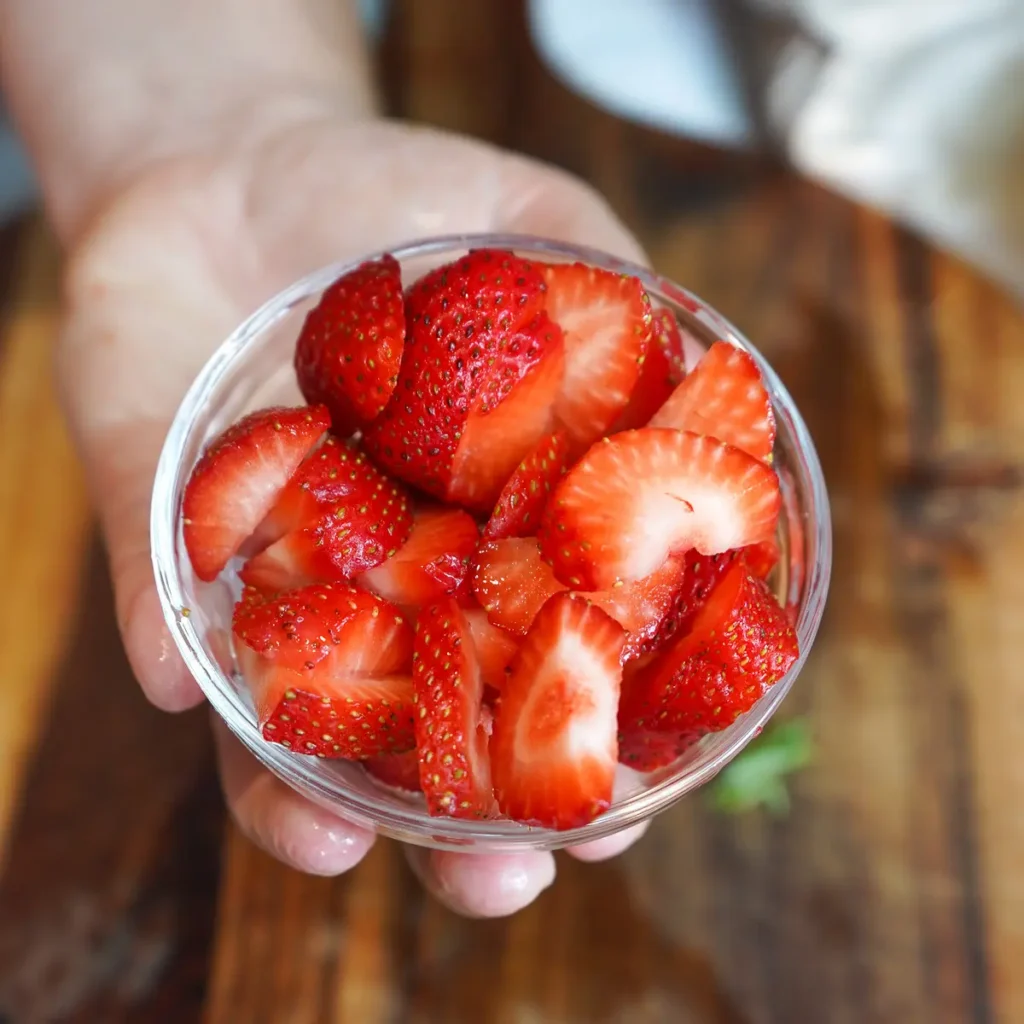 Sliced fresh red strawberries in a small clear glass bowl held in hand.