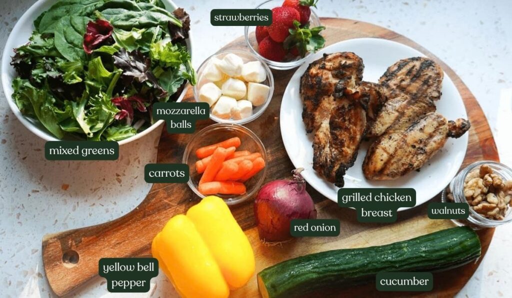 Flat lay of fresh ingredients for a grilled chicken salad including strawberries, cucumbers, bell peppers, walnuts, and mozzarella.