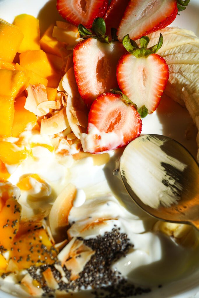 Close-up spoon scooping creamy Greek yogurt with diced mango, crisp toasted coconut flakes, and golden raw honey.
